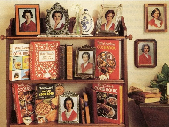 Three tier wood shelf in a kitchen displaying 6 Betty Crocker cookbook editions from 1950-1991, aside framed images of Betty Crocker headshots through the years.